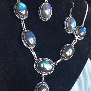 Puede incluir: Un conjunto de collar y pendientes de plata con piedras de labradorita de forma ovalada. Las piedras tienen un brillo iridiscente azul oscuro y verde.