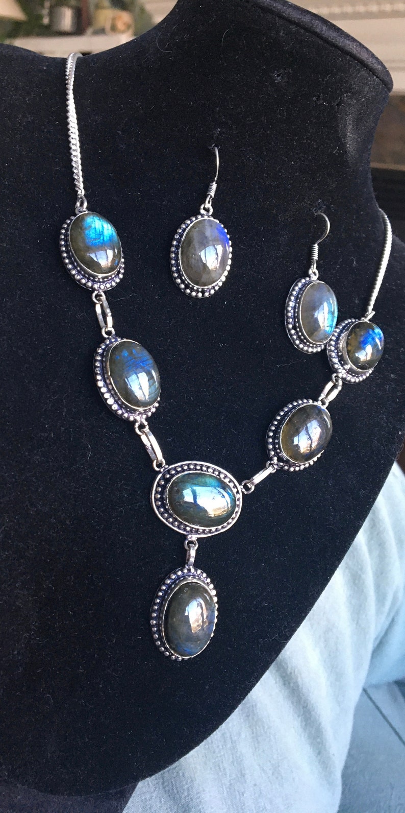 Puede incluir: Un conjunto de collar y pendientes de plata con piedras de labradorita de forma ovalada. Las piedras tienen un brillo iridiscente azul oscuro y verde.