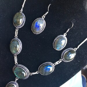 Puede incluir: Un collar y pendientes de plata con piedras de labradorita de forma ovalada. Las piedras tienen una base gris oscura con destellos iridiscentes azules y verdes.