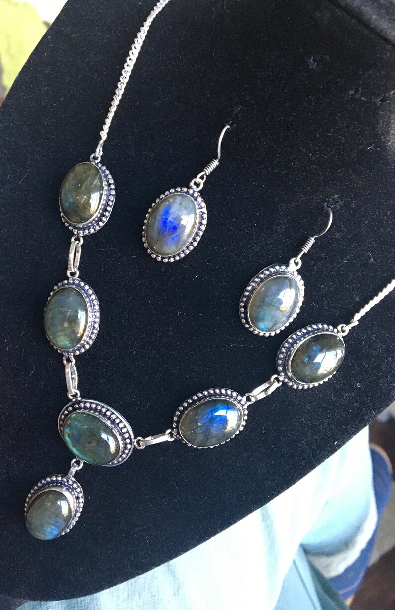 Puede incluir: Un collar y pendientes de plata con piedras de labradorita de forma ovalada. Las piedras tienen una base gris oscura con destellos iridiscentes azules y verdes.