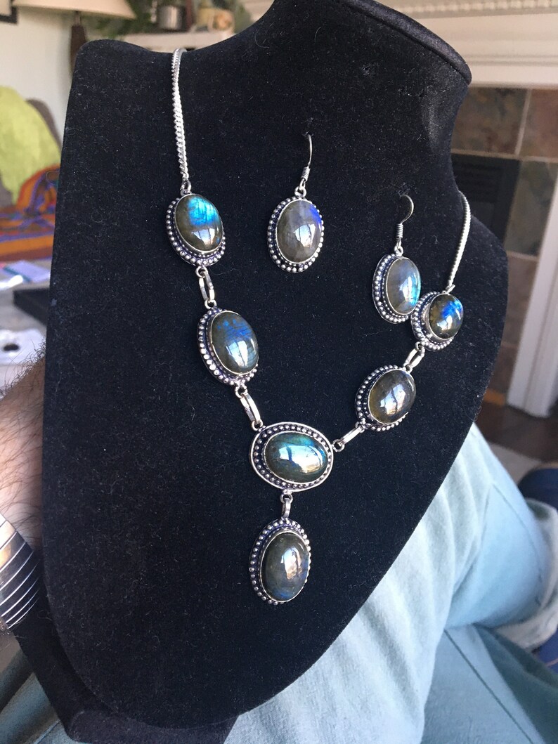 Puede incluir: Un conjunto de collar y pendientes de plata con piedras de labradorita azul iridiscente de forma ovalada. El collar tiene cinco piedras y los pendientes tienen una piedra cada uno.