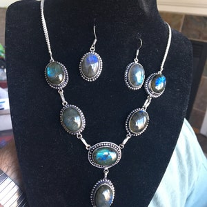 Puede incluir: Un conjunto de collar y pendientes de plata con piedras de labradorita iridiscente de forma ovalada. El collar tiene cinco piedras y un colgante con una piedra m&aacute;s grande. Los pendientes tienen cada uno una piedra.