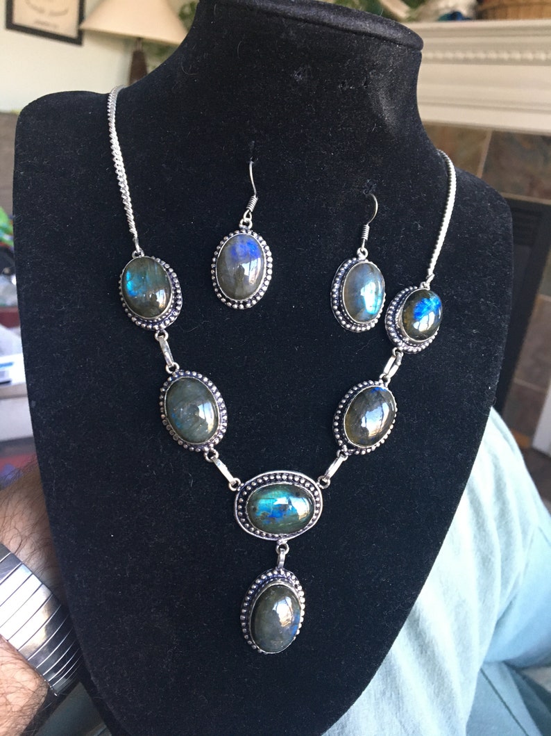 Puede incluir: Un conjunto de collar y pendientes de plata con piedras de labradorita iridiscente de forma ovalada. El collar tiene cinco piedras y un colgante con una piedra m&aacute;s grande. Los pendientes tienen cada uno una piedra.