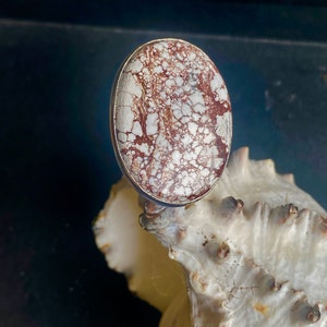 K&ouml;nnte beinhalten: Ein silberner Ring mit einem ovalen Stein in Wei&szlig; und Braun. Der Stein hat ein einzigartiges Muster aus wei&szlig;en und braunen Wirbeln.