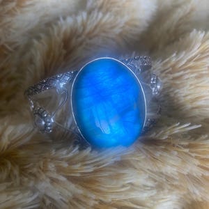 May include: A silver bracelet with a large oval blue gemstone. The bracelet has a delicate design with floral accents.