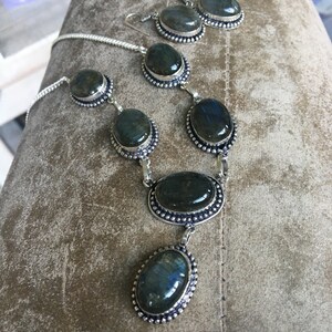 Puede incluir: Un conjunto de collar y pendientes de plata con piedras de labradorita de forma ovalada. Las piedras son de color verde oscuro con destellos iridiscentes azules y dorados.
