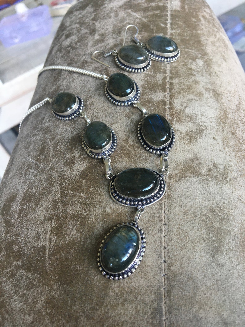 Puede incluir: Un conjunto de collar y pendientes de plata con piedras de labradorita de forma ovalada. Las piedras son de color verde oscuro con destellos iridiscentes azules y dorados.