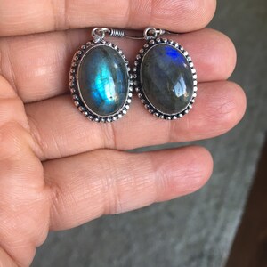 Puede incluir: Un par de pendientes de plata con piedras de labradorita de forma ovalada. Las piedras tienen una base gris oscura con destellos iridiscentes azules y verdes.