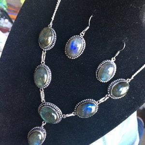 Puede incluir: Un conjunto de collar y pendientes de plata con piedras de labradorita de forma ovalada. Las piedras tienen un brillo iridiscente verde oscuro y azul.