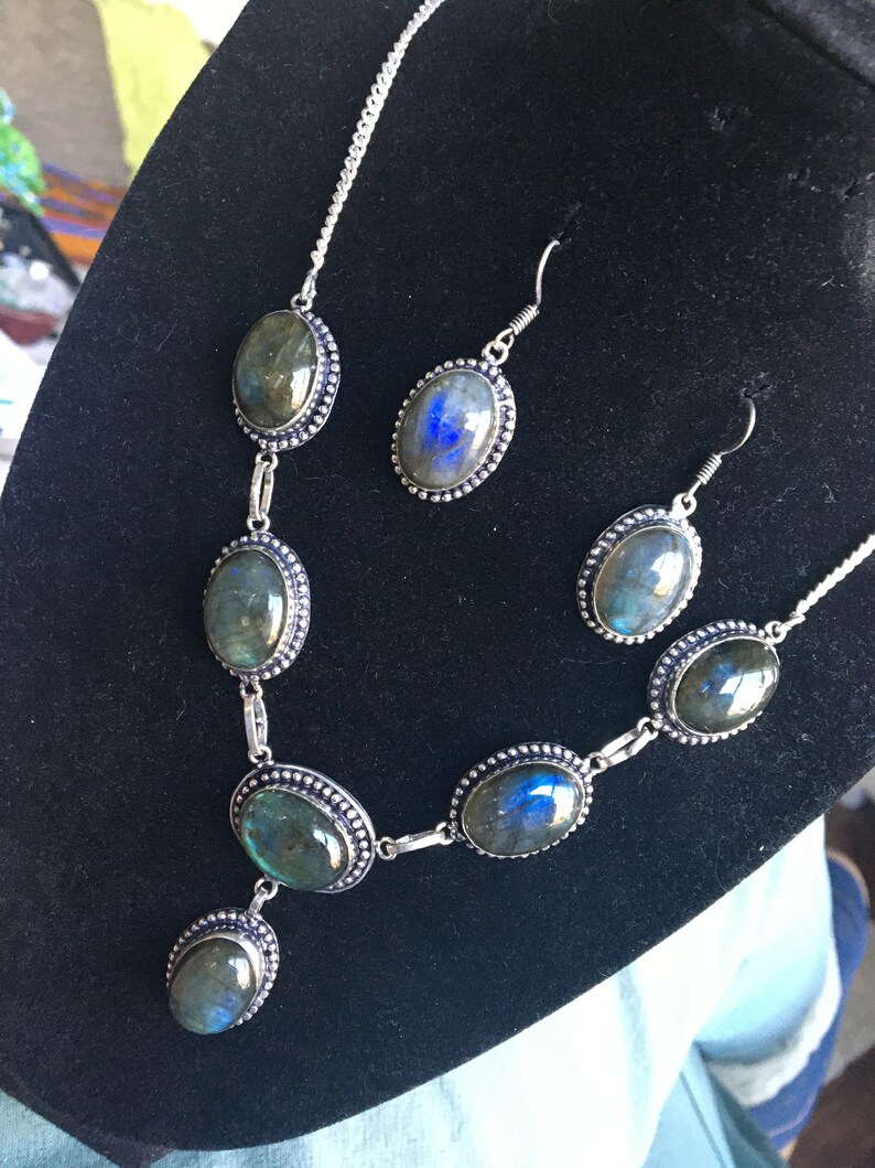 Puede incluir: Un conjunto de collar y pendientes de plata con piedras de labradorita de forma ovalada. Las piedras tienen un brillo iridiscente verde oscuro y azul.