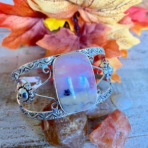 May include: A silver cuff bracelet with a large, rectangular, pink and white stone center. The bracelet has intricate silver designs and is displayed on a wooden surface with colorful autumn leaves and rocks.