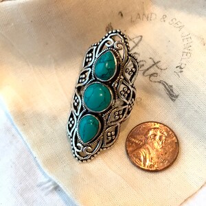 K&ouml;nnte beinhalten: Ein silberner Ring mit einem verzierten Design und drei t&uuml;rkisfarbenen Steinen. Der Ring liegt auf einem wei&szlig;en Stoffhintergrund mit dem Text "Land & Sea Jewelers".