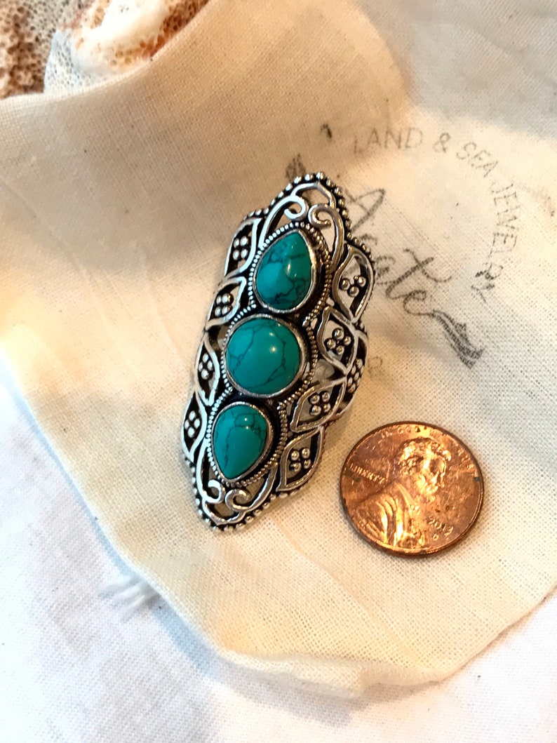 K&ouml;nnte beinhalten: Ein silberner Ring mit einem verzierten Design und drei t&uuml;rkisfarbenen Steinen. Der Ring liegt auf einem wei&szlig;en Stoffhintergrund mit dem Text "Land & Sea Jewelers".