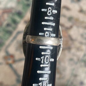 K&ouml;nnte beinhalten: Ein silberner Ring mit einem schwarzen Stein und einem silbernen Band. Der Ring ist auf einem Lineal mit der Gr&ouml;&szlig;e 10 gemessen.