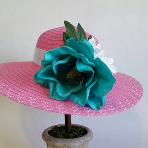 May include: A pink sun hat with a white ribbon band and a large turquoise flower. The hat has a wide brim with white polka dots. The flower is the focal point, with green leaves and white flowers accenting the design. The hat is displayed on a decorative stand.