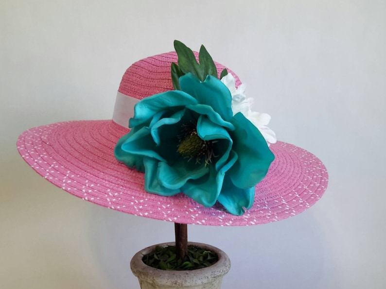 May include: A pink sun hat with a white ribbon band and a large turquoise flower. The hat has a wide brim with white polka dots. The flower is the focal point, with green leaves and white flowers accenting the design. The hat is displayed on a decorative stand.