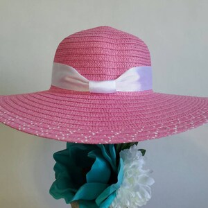 May include: A pink sun hat with a wide brim and a white ribbon bow. The hat has a woven texture and small white dots along the edge of the brim. The hat is a stylish accessory for sunny days.