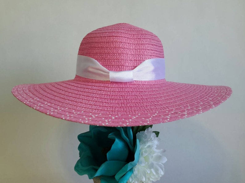May include: A pink sun hat with a wide brim and a white ribbon bow. The hat has a woven texture and small white dots along the edge of the brim. The hat is a stylish accessory for sunny days.