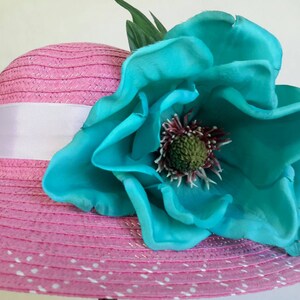 May include: A pink sun hat with a white ribbon and a large turquoise flower. The hat has a wide brim with white polka dots. The flower has a detailed center and green leaves.