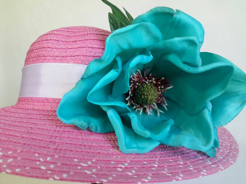 May include: A pink sun hat with a white ribbon and a large turquoise flower. The hat has a wide brim with white polka dots. The flower has a detailed center and green leaves.