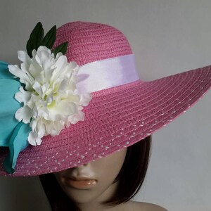 May include: A pink sun hat with a wide brim, decorated with a white ribbon and large floral accents in white, teal, and green. The hat has a dotted pattern and is designed for sun protection.
