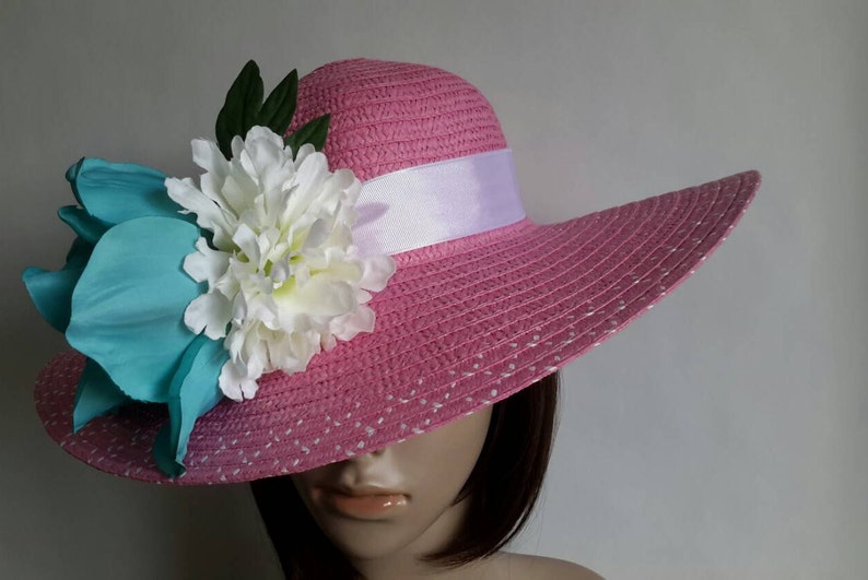 May include: A pink sun hat with a wide brim, decorated with a white ribbon and large floral accents in white, teal, and green. The hat has a dotted pattern and is designed for sun protection.