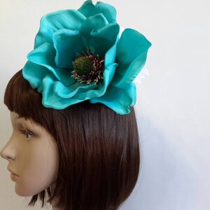 May include: A large, teal-colored flower hair accessory. The flower has multiple layers of petals and a detailed center with green and brown accents. The accessory is attached to a hair clip or comb, designed to be worn in the hair.