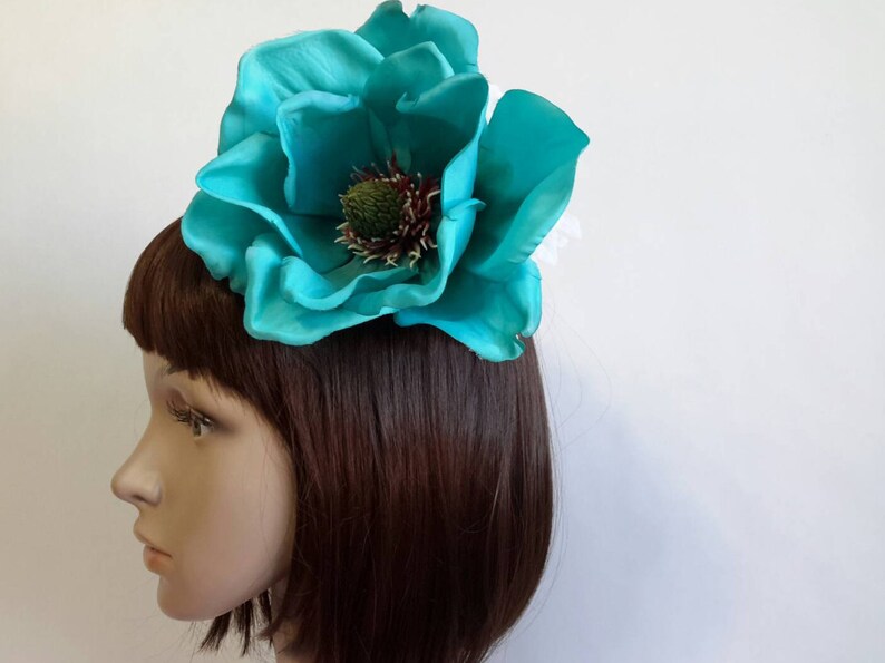 May include: A large, teal-colored flower hair accessory. The flower has multiple layers of petals and a detailed center with green and brown accents. The accessory is attached to a hair clip or comb, designed to be worn in the hair.