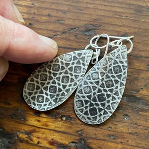 Könnte beinhalten: Ein Paar silberne Tropfenohrringe mit einem strukturierten, geometrischen Muster. Die Ohrringe haben einen Hakenverschluss und werden auf einer Holzoberfläche präsentiert. Das Muster ist eine sich wiederholende Reihe kleiner Quadrate und Formen.