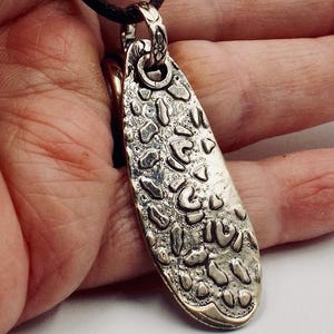 May include: Silver-toned pendant with a textured surface and a dark brown cord. The pendant has an oval shape with an irregular pattern of raised shapes. The cord is attached to a decorative bail at the top.
