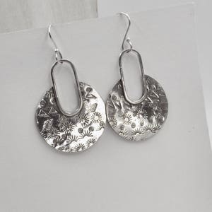 Peut inclure: Une paire de boucles d'oreilles en argent avec un motif circulaire. Les boucles d'oreilles ont une surface texturée avec un motif de petits cercles et triangles. Les boucles d'oreilles sont suspendues à un crochet en argent.