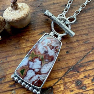 May include: A silver necklace with a toggle clasp and a pendant featuring a rectangular piece of agate with brown, white, and green swirls.