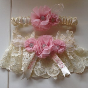 May include: Two ivory lace garters with pink satin bows and pink flowers. The larger garter has a satin ribbon with the text "May 12/16".