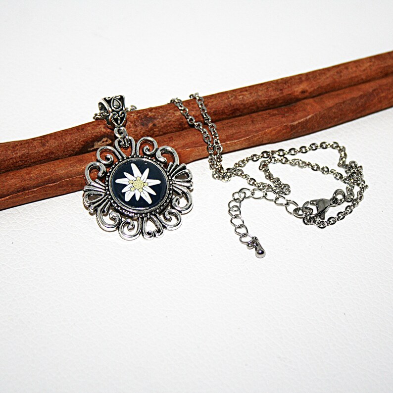 May include: A silver necklace with a pendant featuring a white edelweiss flower on a black background. The pendant is surrounded by a decorative silver frame. The necklace is on a brown wooden surface.