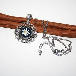May include: A silver necklace with a silver pendant featuring a white edelweiss flower on a black background. The pendant is ornate and has a delicate chain.