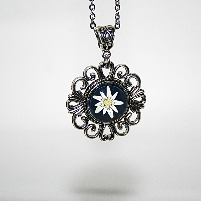 May include: A silver necklace with a pendant featuring a white edelweiss flower on a dark blue background. The pendant is surrounded by a decorative silver frame.