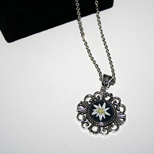 May include: A silver necklace with a round pendant featuring a white edelweiss flower on a black background. The pendant is surrounded by a silver filigree design.