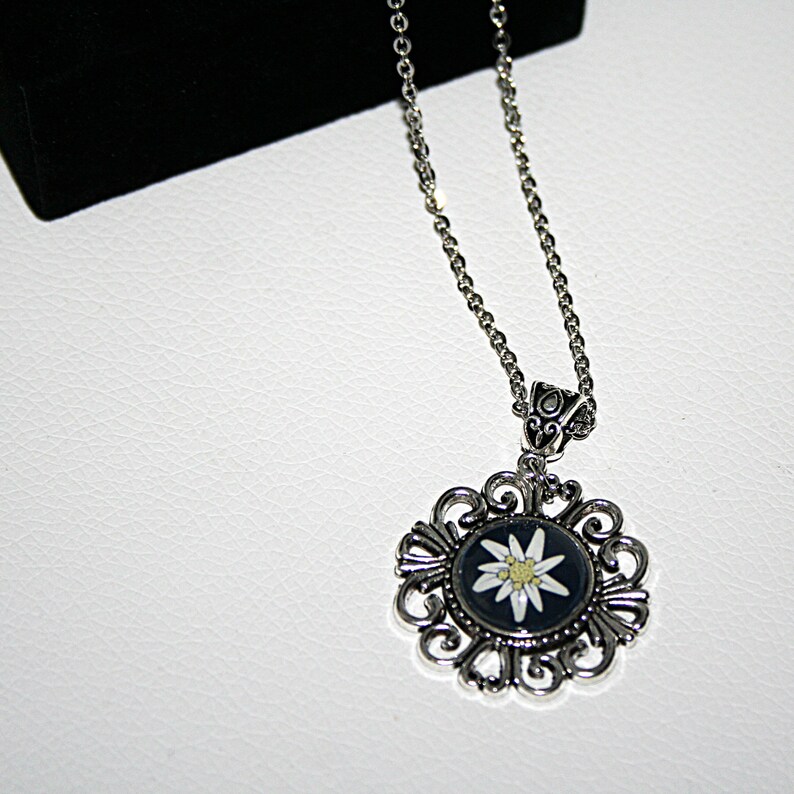 May include: A silver necklace with a round pendant featuring a white edelweiss flower on a black background. The pendant is surrounded by a silver filigree design.
