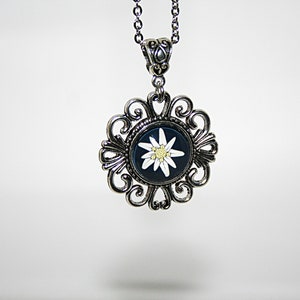 May include: A silver necklace with a pendant featuring a white edelweiss flower on a dark blue background. The pendant is surrounded by a decorative silver frame.