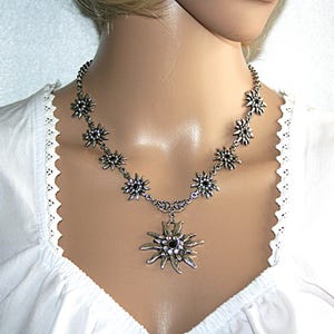May include: Silver-coloured necklace with a floral design and black and clear crystal accents. The necklace has a pendant with a larger flower design. The necklace is worn over a white blouse.