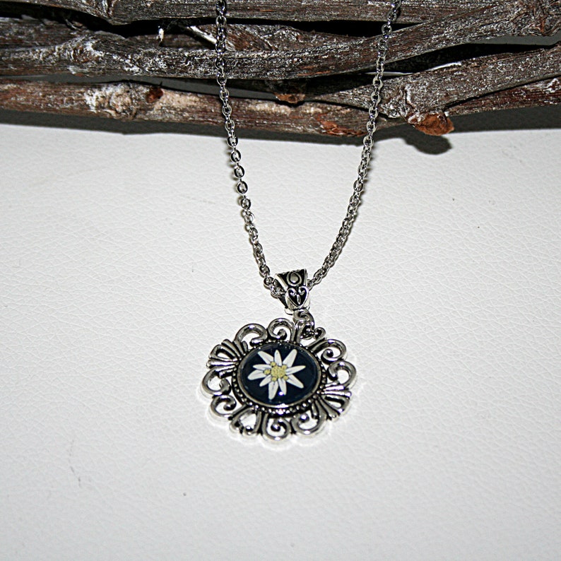 May include: A silver chain necklace with a pendant featuring a white edelweiss flower on a black background. The pendant is surrounded by a silver filigree design.