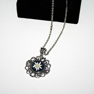 May include: A silver necklace with a round pendant featuring a white edelweiss flower on a black background. The pendant is surrounded by an intricate silver filigree design.