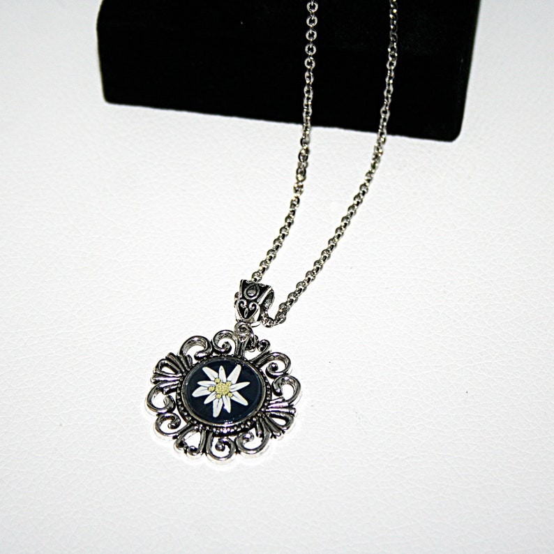 May include: A silver necklace with a round pendant featuring a white edelweiss flower on a black background. The pendant is surrounded by an intricate silver filigree design.