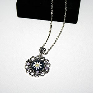 May include: A silver necklace with a pendant featuring a white edelweiss flower on a black background. The pendant is surrounded by a silver filigree design.