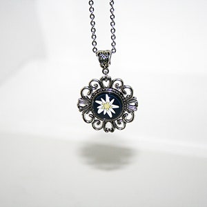 May include: A silver necklace with a pendant featuring a white edelweiss flower on a black background. The pendant is surrounded by a decorative silver frame.