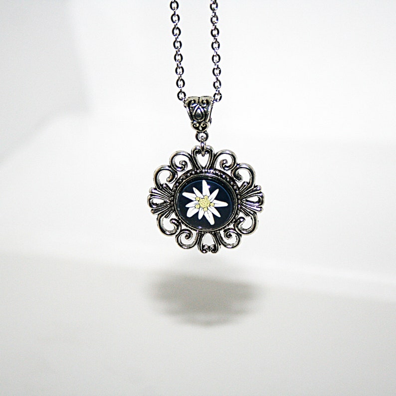 May include: A silver necklace with a pendant featuring a white edelweiss flower on a black background. The pendant is surrounded by a decorative silver frame.