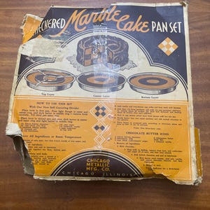Op de afbeelding: Vintage "Checkered Marble Cake Pan Set" doos. De doos bevat illustraties van een marmercake en de onderdelen van de pannenset. Instructies en een recept voor chocoladeboterglazuur zijn inbegrepen. Het logo van Chicago Metallic Mfg. Co. is zichtbaar.