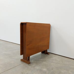 May include: A rectangular wooden drop-leaf table in a medium brown finish, shown in its fully folded, upright position. The table features a smooth, flat surface on the side and is supported by two vertical legs with horizontal feet at the base.