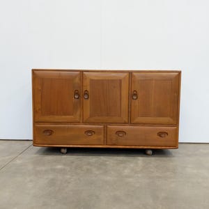 May include: A wooden cabinet with three doors and two drawers. The cabinet is made of light brown wood and has a rectangular shape. The doors have round handles, and the drawers have oval handles. The cabinet rests on small, round feet.