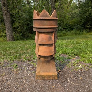 May include: A tall, weathered terracotta chimney pot featuring a crown-shaped top with pointed edges. The cylindrical body displays a series of stacked, flared tiers with vertical support fins, finished in a rustic, earthy orange-brown texture.